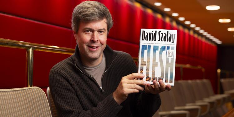 Author David Szalay sits smiling and holding up his book, Flesh